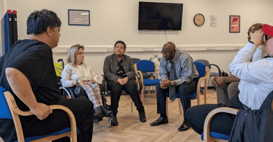 A diverse group of six people sit in a circle having a discussion in a room with chairs, a clock, and a TV on the wall.