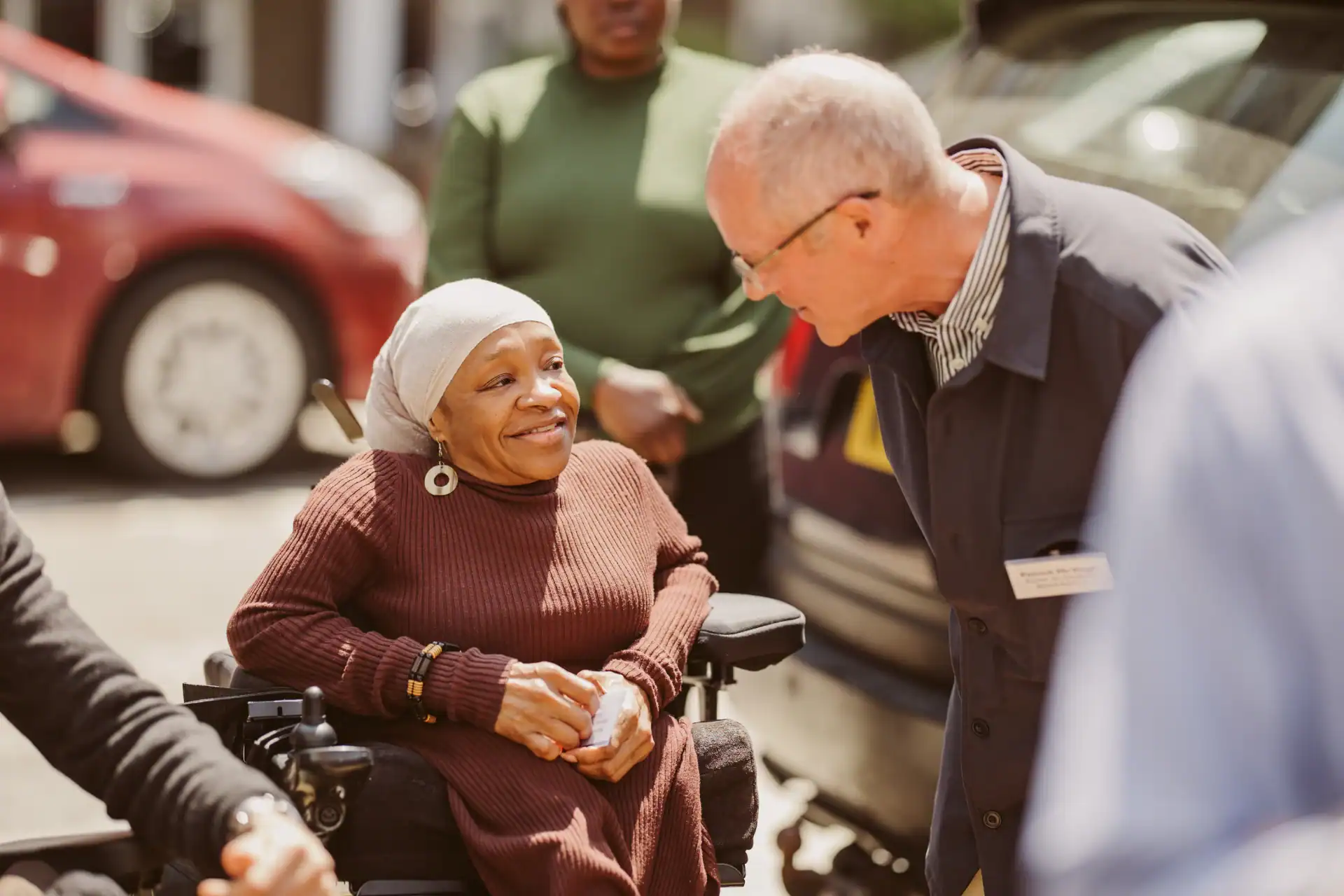 A woman in a wheelchair smiles and talks with an older man outdoors, with two other people and parked cars in the background.