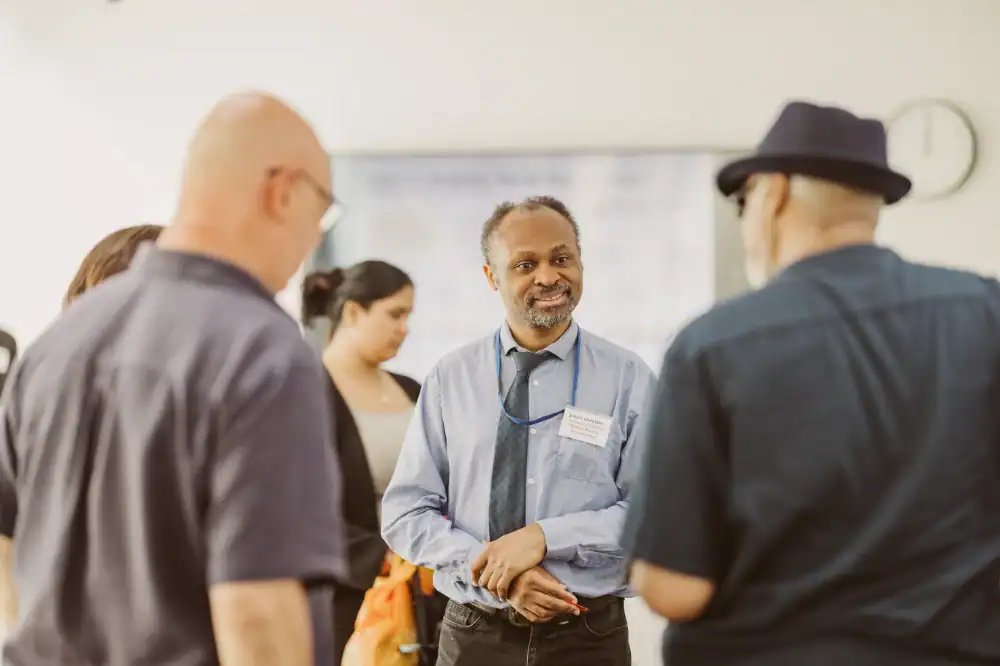 A man with a name badge and tie talks to three people in a brightly lit room; others and a clock are visible in the background.