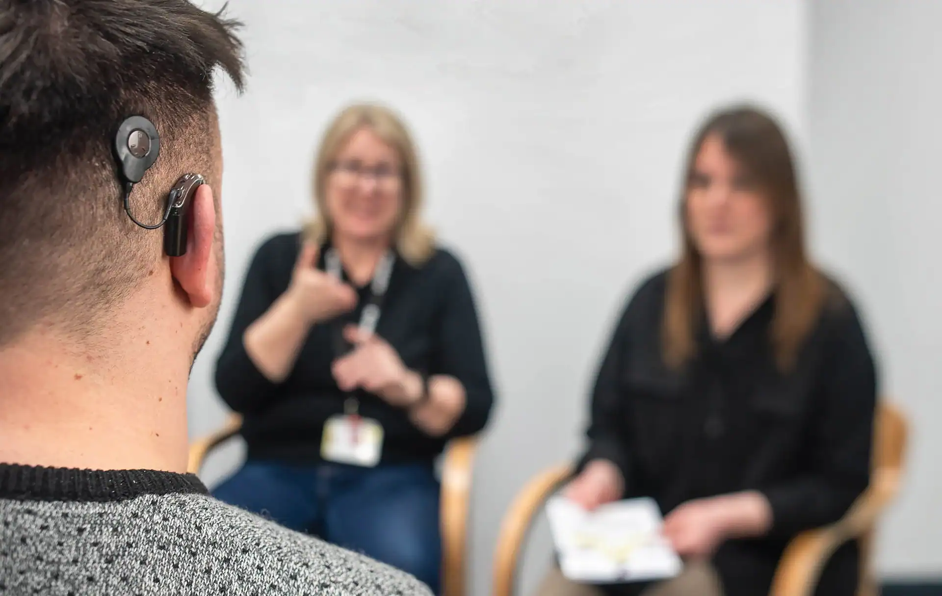 A person with a cochlear implant sits facing two women; one is using sign language and the other is holding papers.
