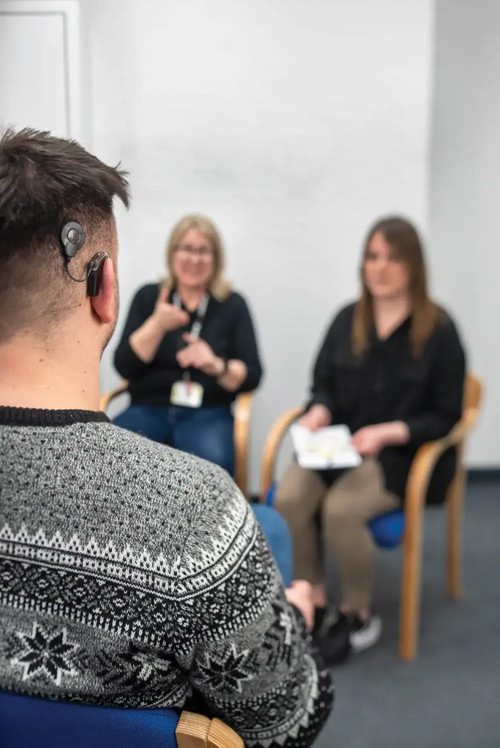 A man with a cochlear implant sits facing two women; one woman is using sign language, and the other is holding some papers.