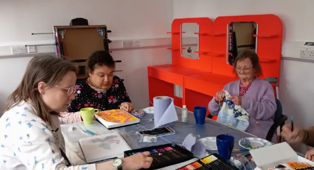 Three women sit around a table doing arts and crafts, with colored pencils, sketchbooks, mugs, and art supplies on the table. Bright orange shelves are in the background.