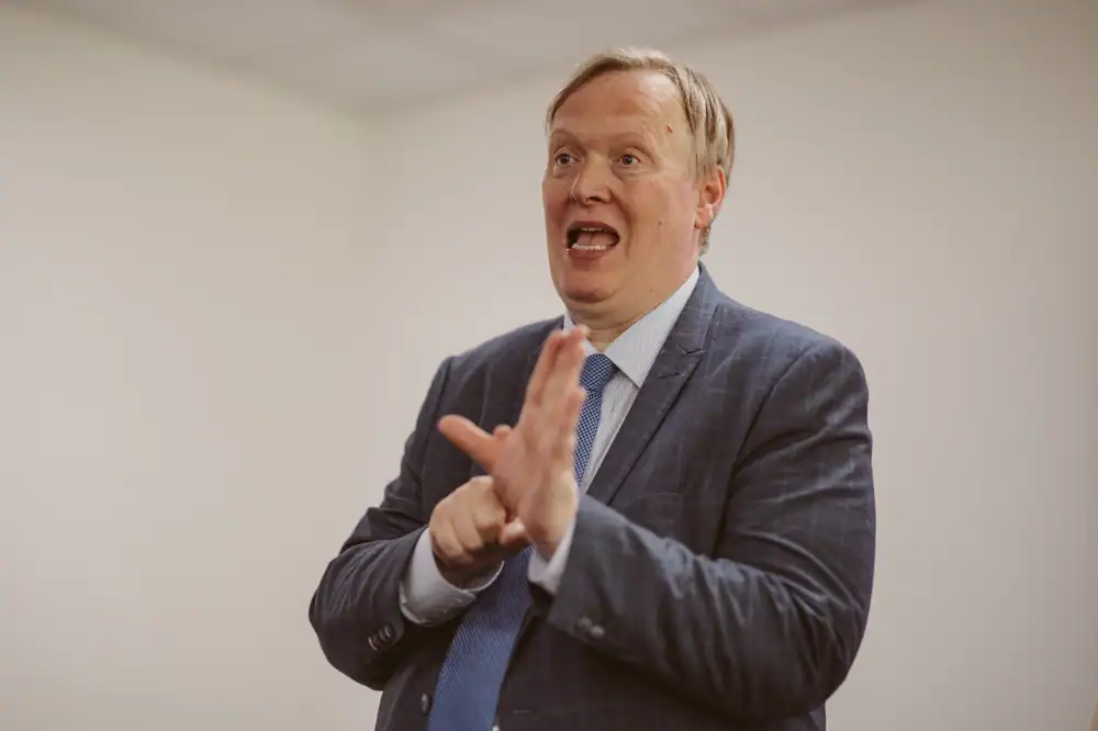 A man in a suit and tie is standing in a room, gesturing with his hands while speaking.