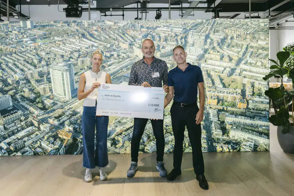 Three people stand indoors holding a large ceremonial check for £10,000, with an aerial cityscape photo displayed on the wall behind them.