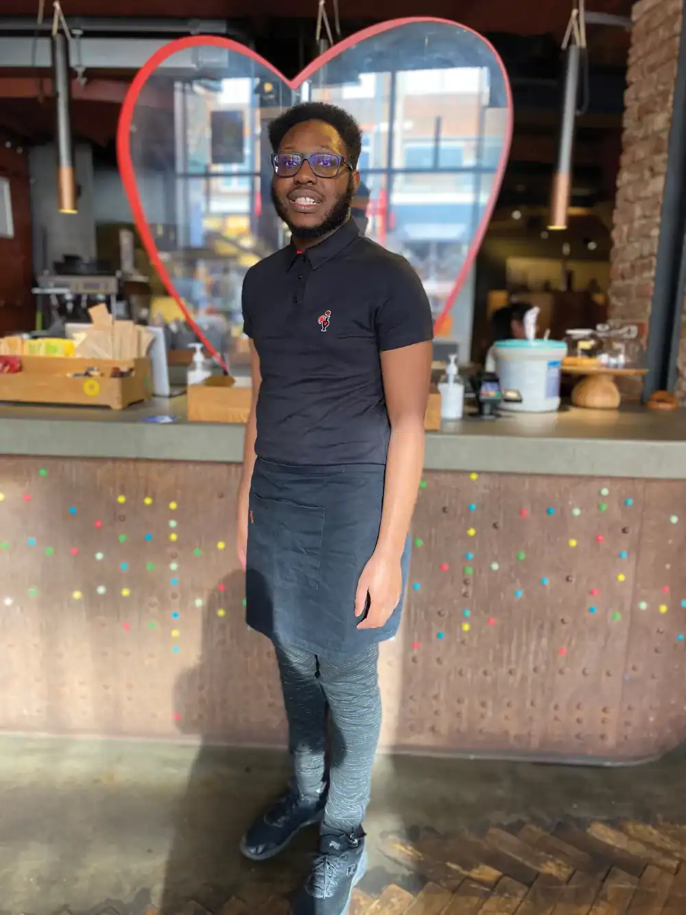 A person wearing glasses, a black polo shirt, and an apron stands in front of a counter with colorful dots and a large red heart outline in the background.