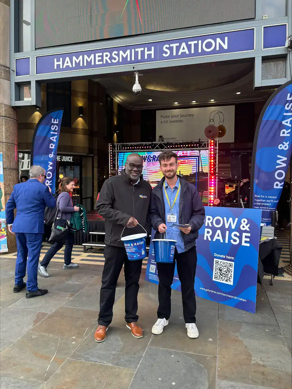 Two people stand outside Hammersmith Station holding donation buckets next to a "Row & Raise" fundraising sign and QR code.