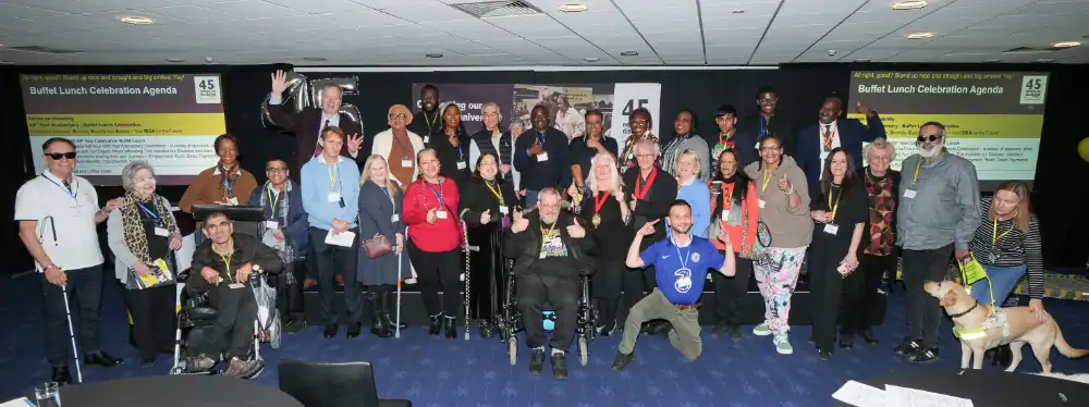 A diverse group of people, including individuals with disabilities and a guide dog, pose together at an indoor event with presentation screens in the background.
