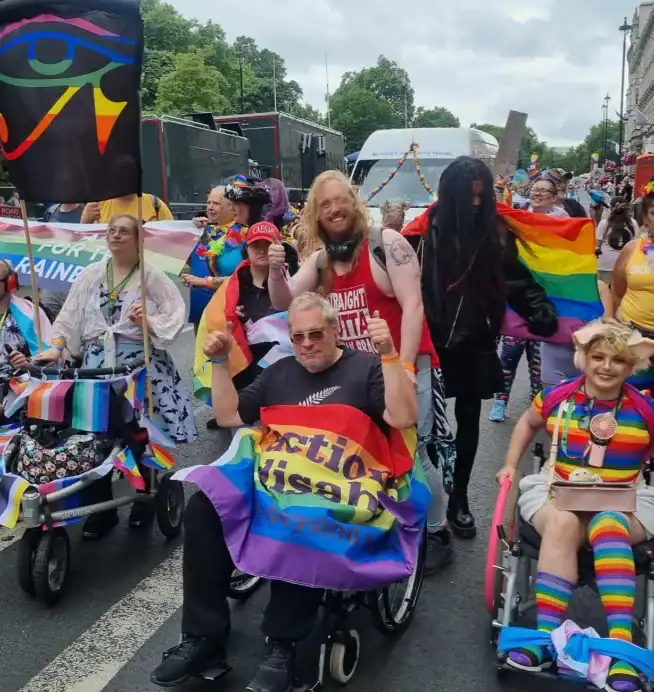 A group of people, some in wheelchairs, participate in a parade holding and wearing various pride flags and signs advocating for disability rights.