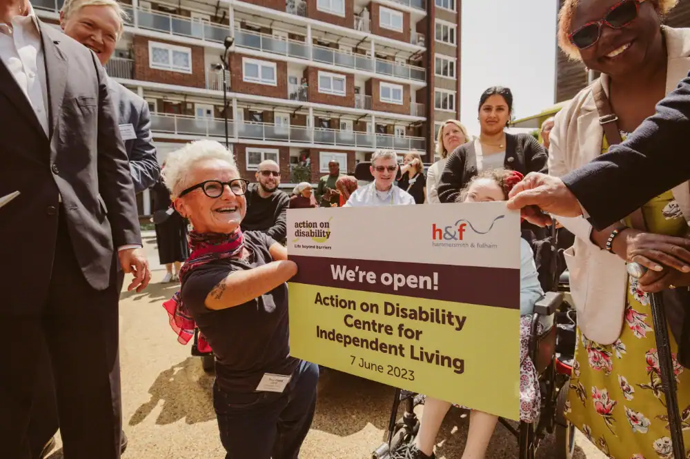 A group of people, including a woman with a limb difference, hold a sign reading “We’re open! Action on Disability Centre for Independent Living, 7 June 2023” at an outdoor event.