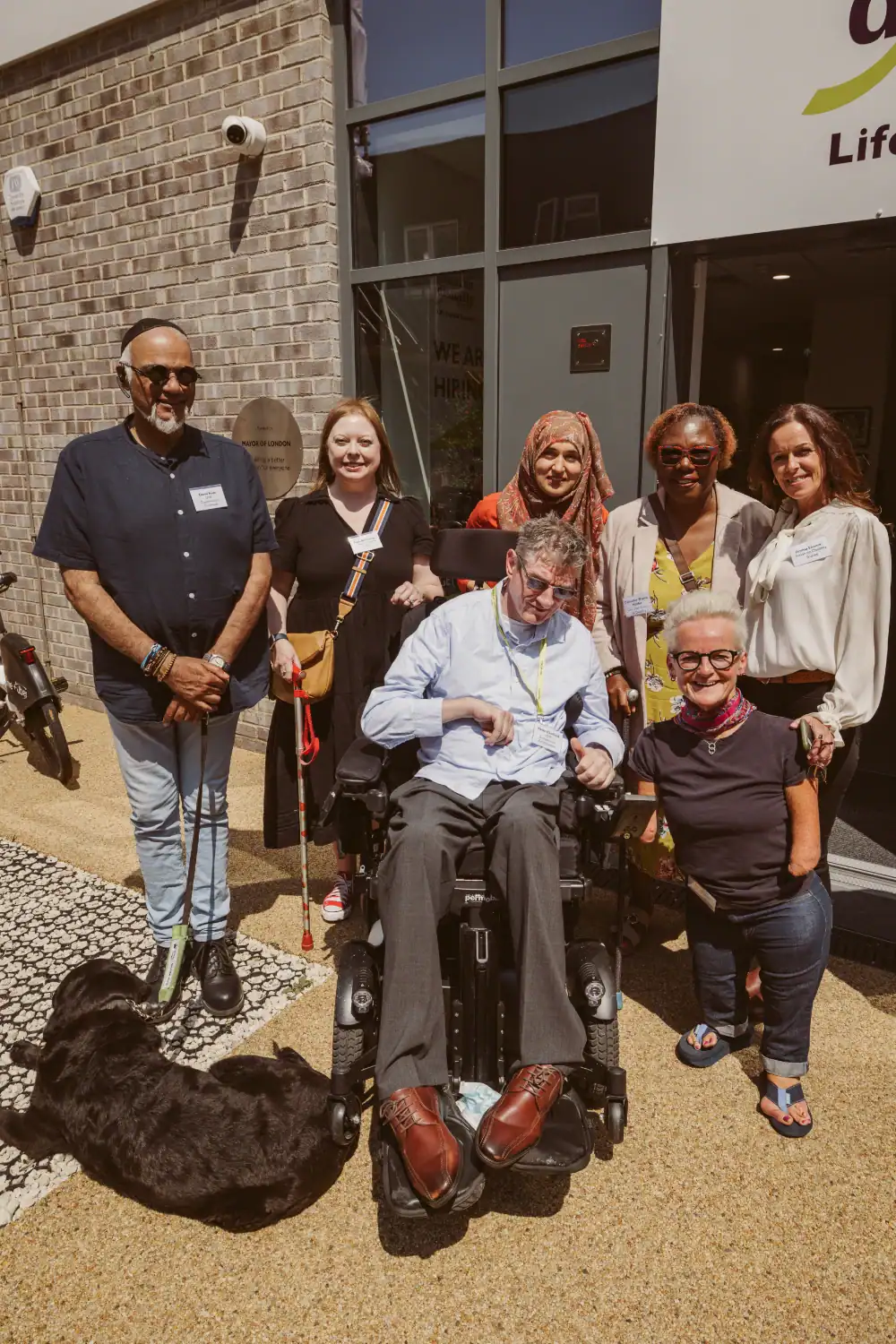 A group of seven adults, including a man in a power wheelchair and a guide dog, pose together outside a building on a sunny day.