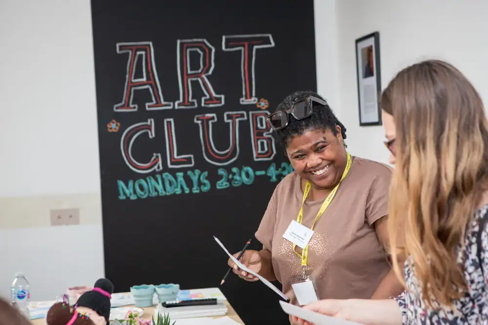 Two women stand in front of a sign that says "Art Club Mondays 2:30-4:30," smiling and holding papers during an indoor art club event.