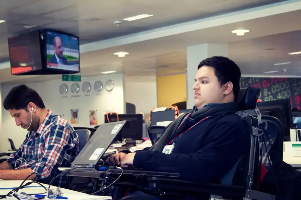 Two men working at desks in an office; one uses a wheelchair and a laptop mounted on an adjustable arm, while the other wears earphones and types on a laptop.