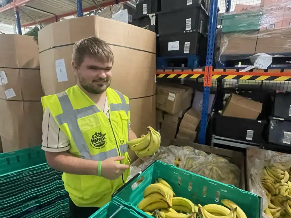 A person wearing a yellow safety vest stands in a warehouse, holding a bunch of bananas near a green crate filled with bananas. Large boxes and plastic bins are stacked in the background.