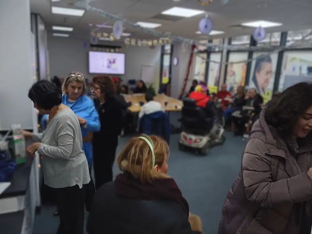 A group of people gather and interact in a brightly lit community room with tables, chairs, and decorations. Some are seated while others stand and talk.