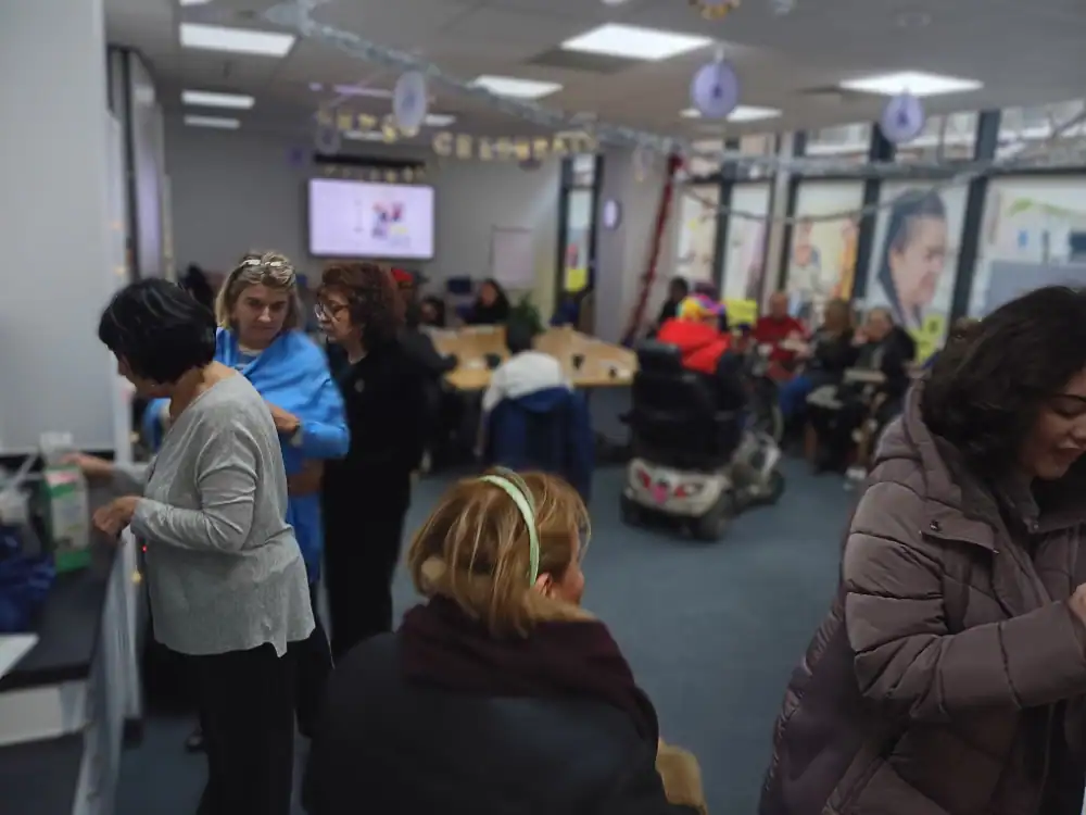 A group of people gather in a brightly lit room with large windows, some seated and some standing near tables, engaged in conversation and activities.