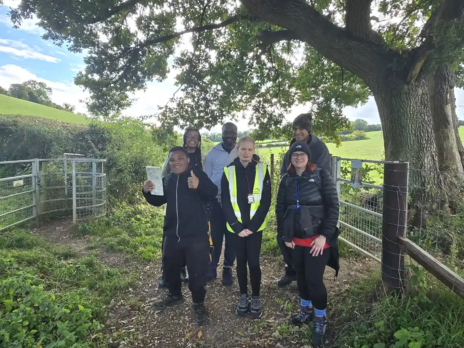 Six people stand under a large tree near a gate in a rural area, with fields in the background. One person holds a map and another wears a reflective vest.
