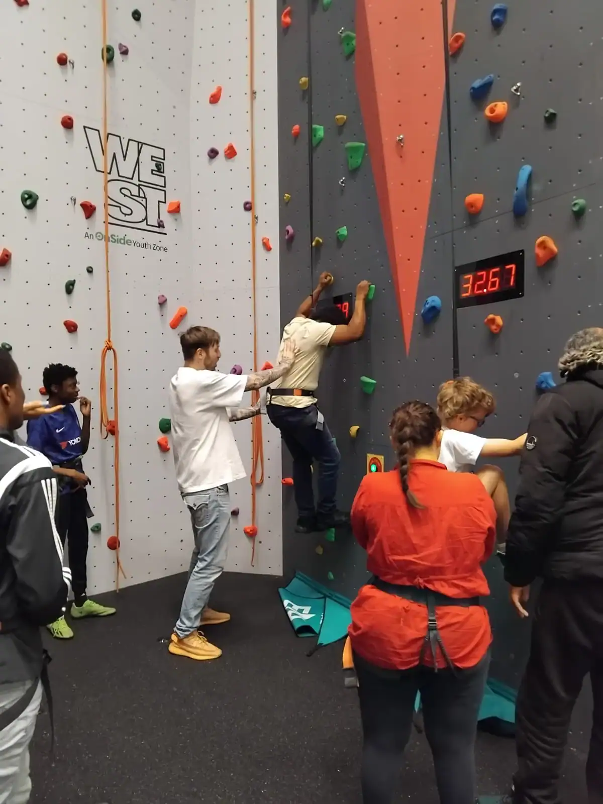 Several people stand around a person climbing an indoor rock wall, while another person assists them. A digital timer on the wall shows 32.67 seconds.