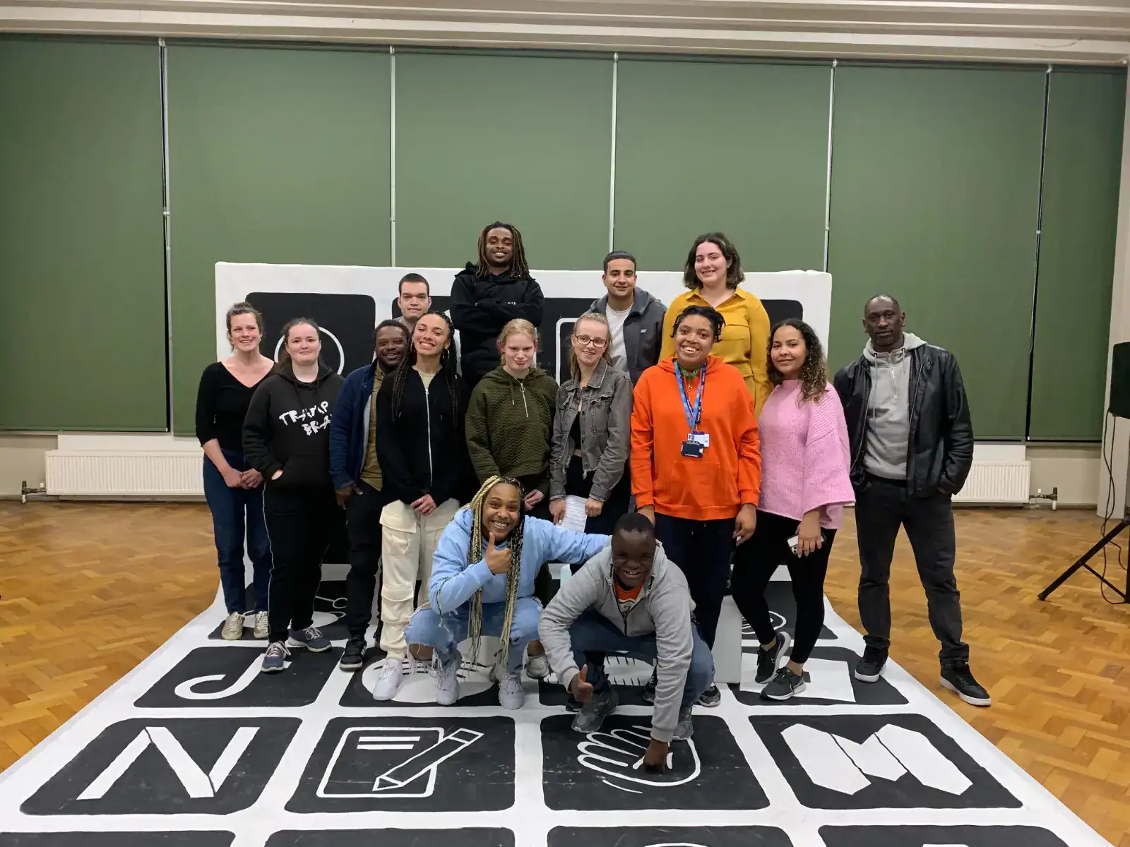 A group of fifteen people pose together indoors, standing and crouching on a large floor graphic with various icons and letters.