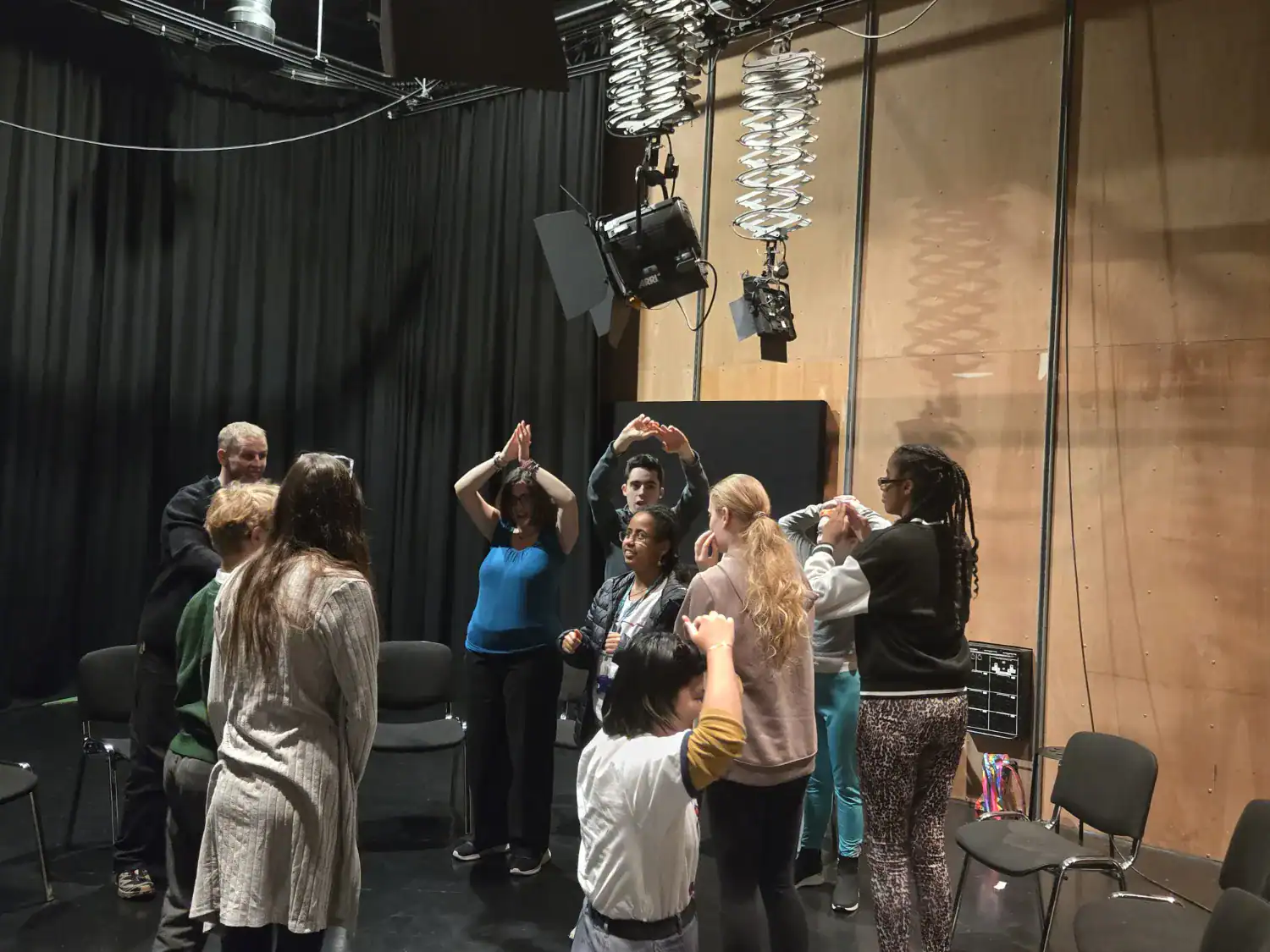 A group of people stand in a circle in a studio, some raising their arms, with chairs arranged around them and lighting equipment visible above.