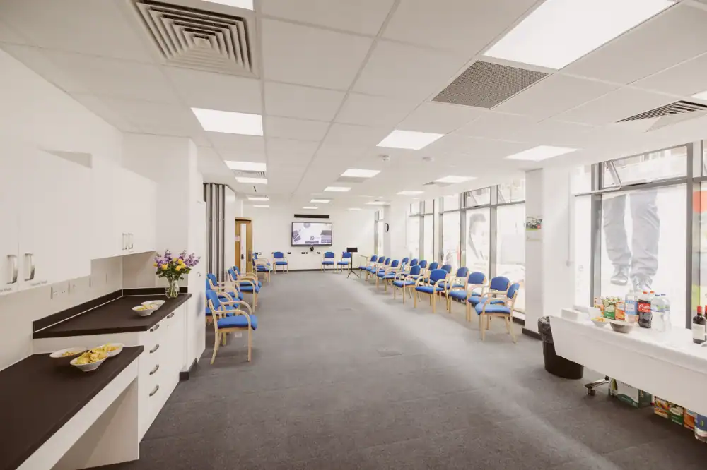 A bright, modern waiting room with rows of blue chairs, a counter with snacks and drinks, large windows, and a flatscreen TV on the back wall.