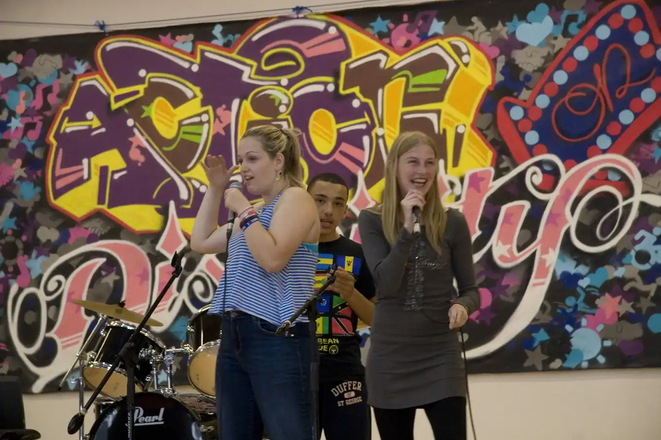 Three young people hold microphones and perform in front of a graffiti-style mural. A drum set is visible in the background.
