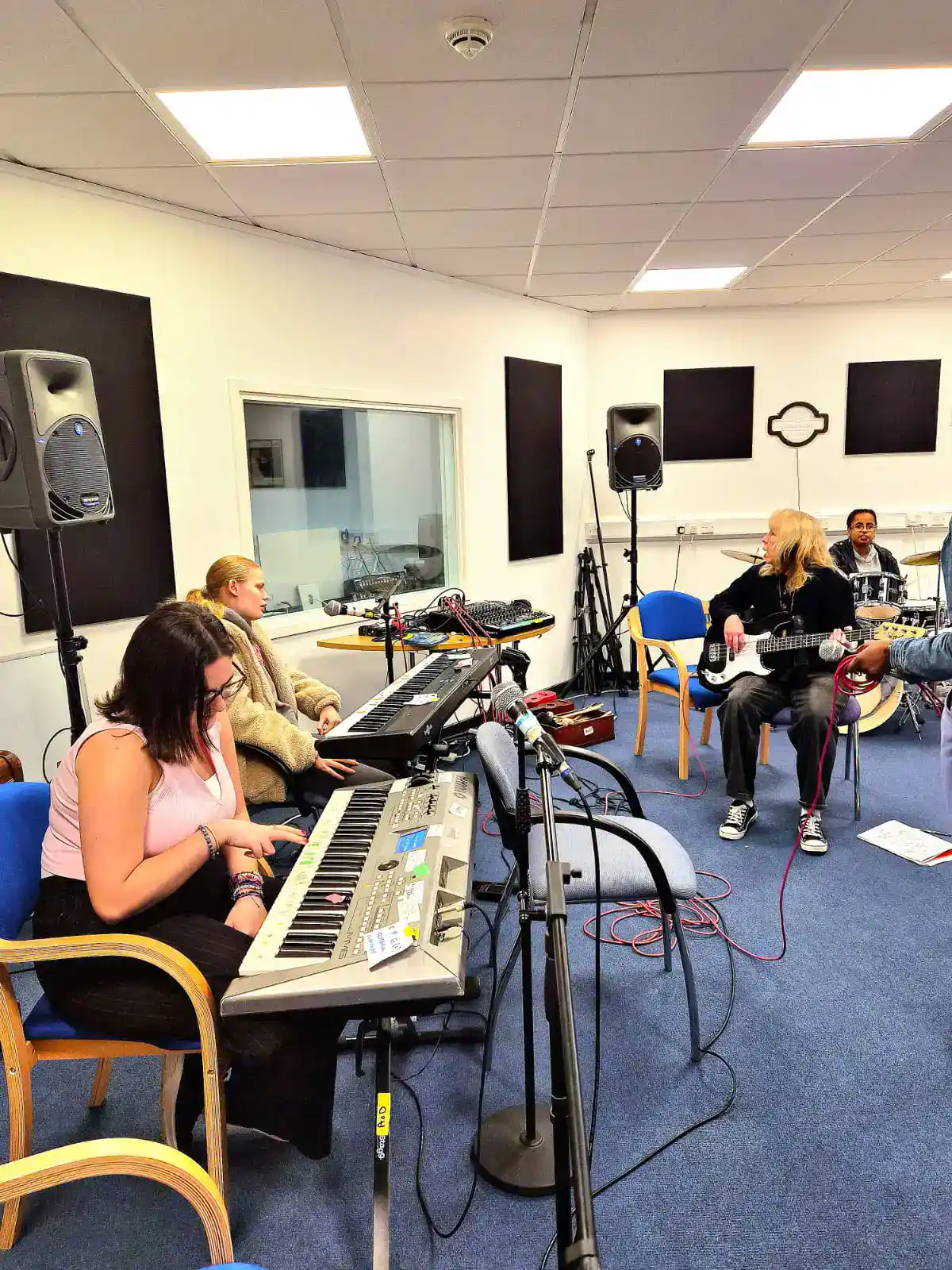A group of people are playing musical instruments, including a keyboard and guitar, in a music rehearsal room with chairs and audio equipment.