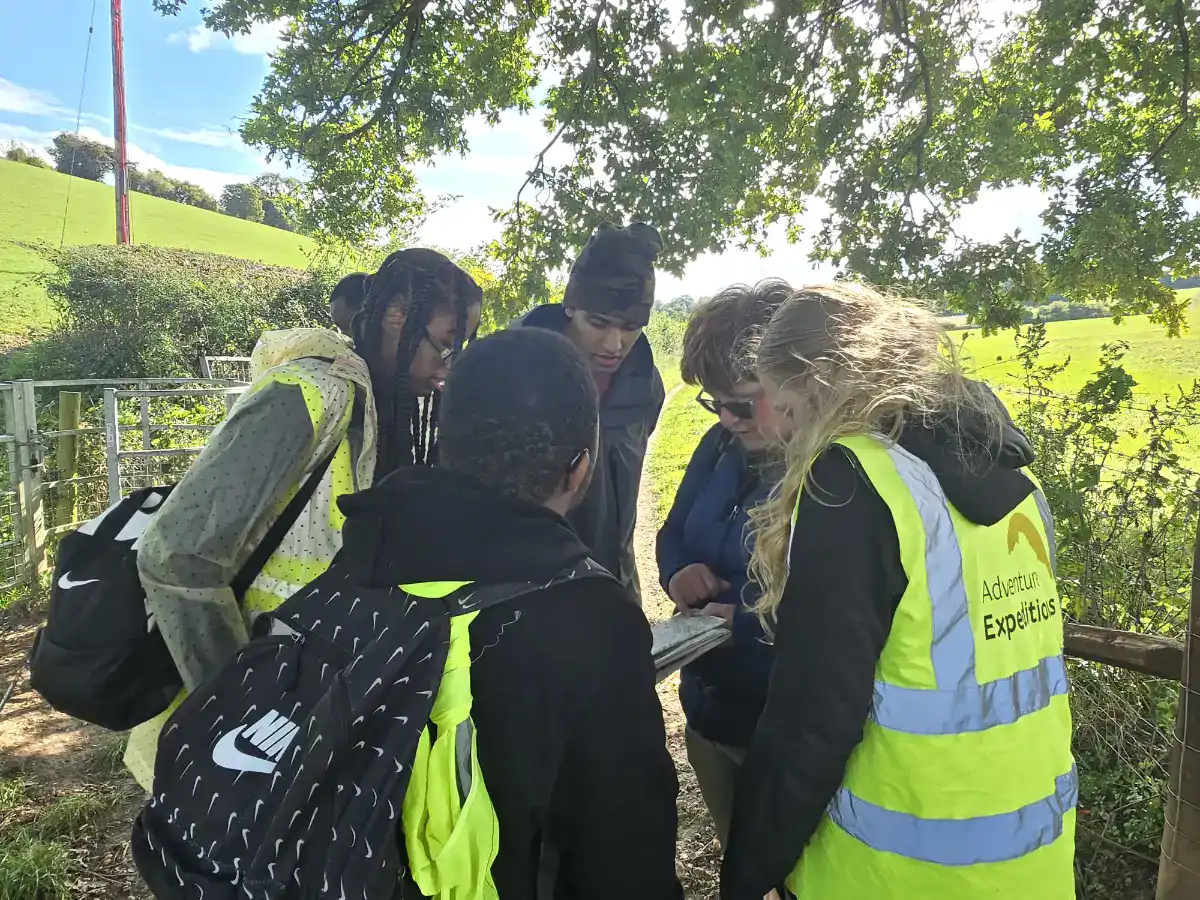 A group of people wearing outdoor gear and high-visibility vests huddle together reading a map in a countryside setting with trees and fields in the background.