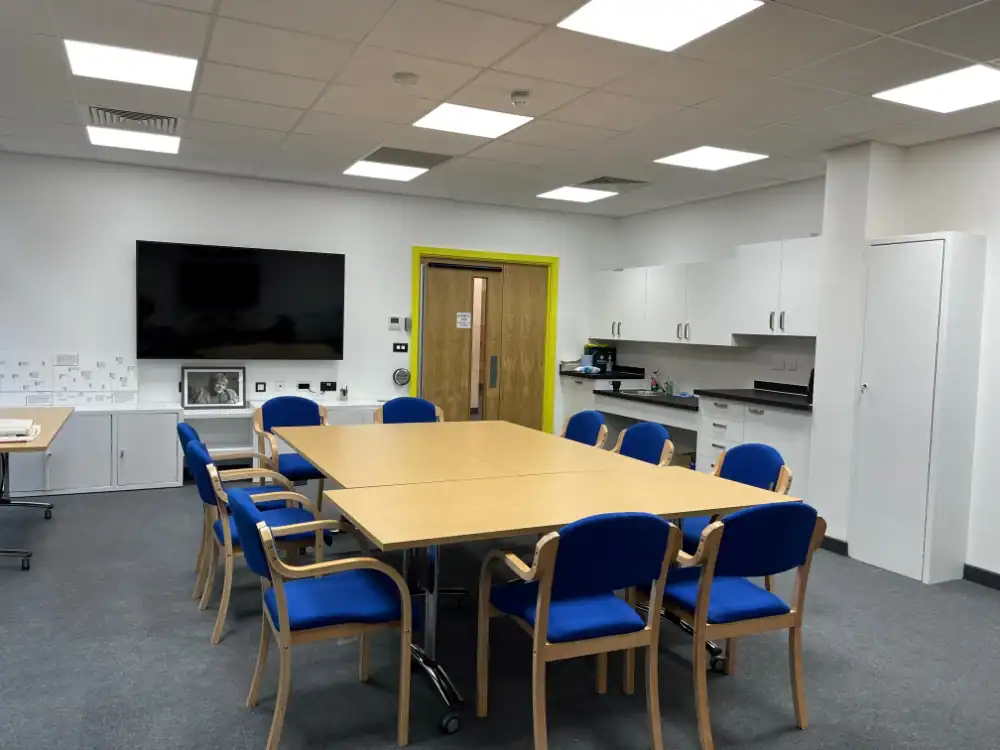 A modern meeting room with a large table, eight blue chairs, a wall-mounted TV, white cabinets, and a wooden door. The room is well-lit with ceiling lights.