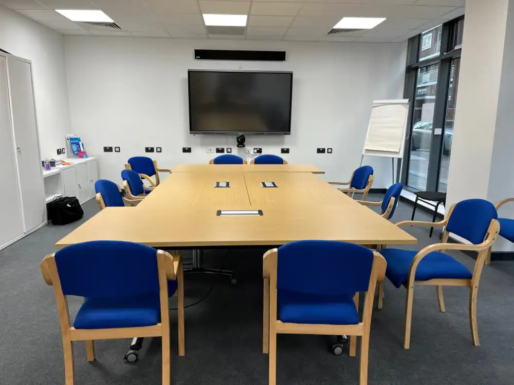 A conference room with a long rectangular table, blue chairs, a large screen on the wall, a flip chart, and windows along one side.