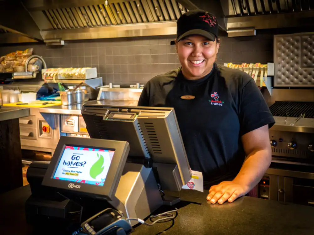 A restaurant employee in uniform stands behind a counter next to a register and kitchen equipment, smiling at the camera.