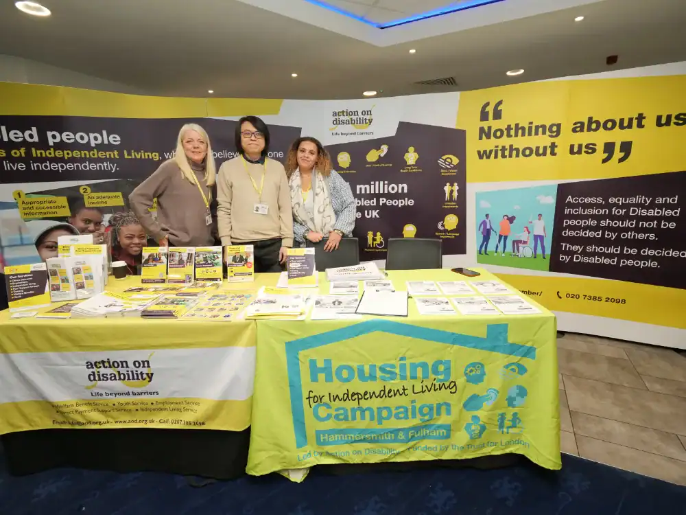 Four people stand behind an information table for Action on Disability and the Housing for Independent Living Campaign, with pamphlets and banners about disability rights on display.