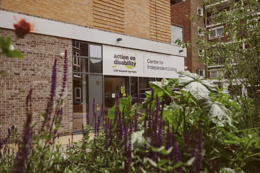 A building entrance with a sign reading "Action on Disability: Centre for Independent Living," partially obscured by green plants and purple flowers in the foreground.
