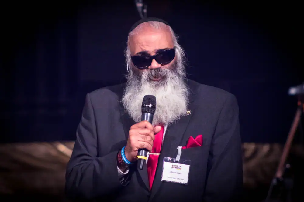 A man with a long gray beard and sunglasses speaks into a microphone while wearing a black suit, red tie, and name badge.