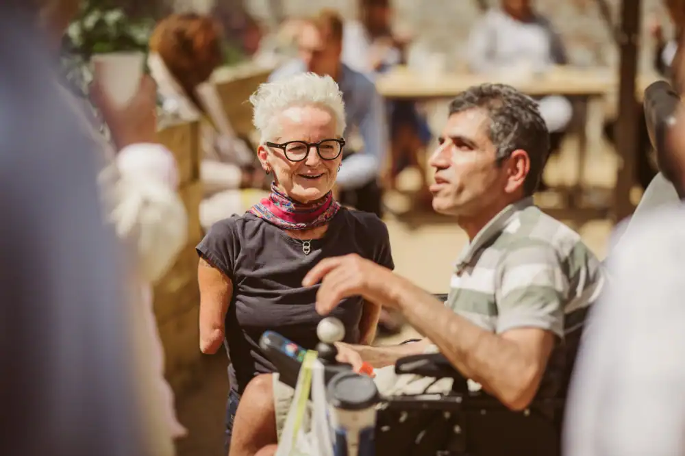 Two people, one woman with a limb difference and a man in a wheelchair, are talking outdoors in a sunny, social setting with others in the background.