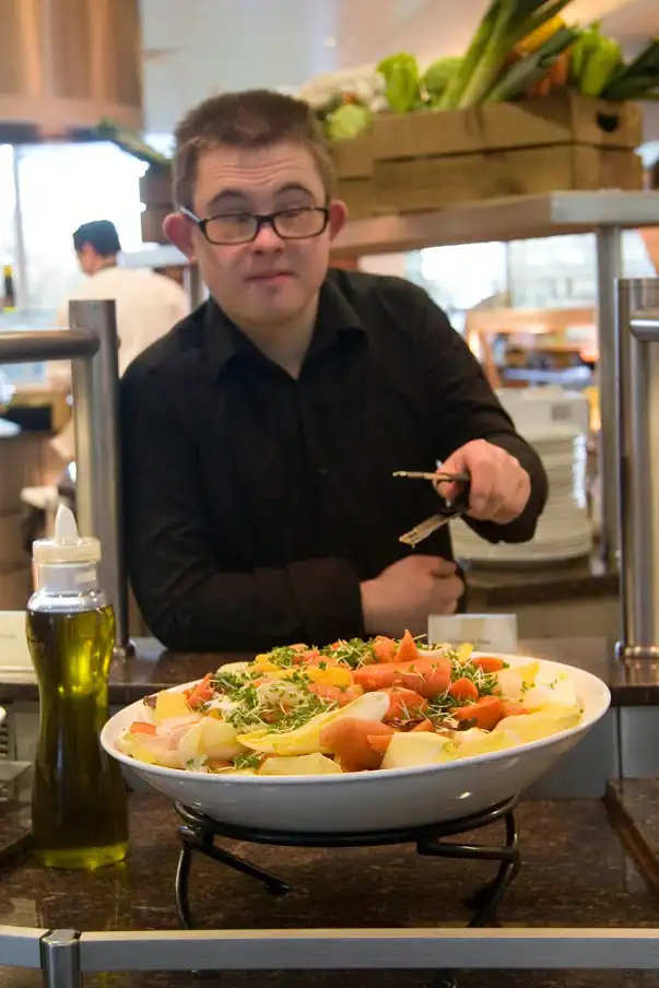 A person wearing glasses and a black shirt serves salad with tongs at a food counter, with a bottle of oil and various vegetables in the background.