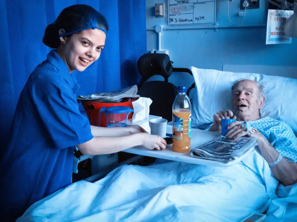 A nurse in blue scrubs serves a drink to an elderly male patient lying in a hospital bed, who holds a cup and a newspaper.