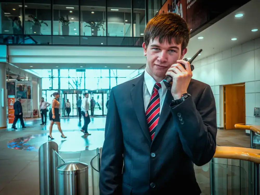 A young man in a suit and striped tie holds a walkie-talkie in a modern office lobby with people walking in the background.