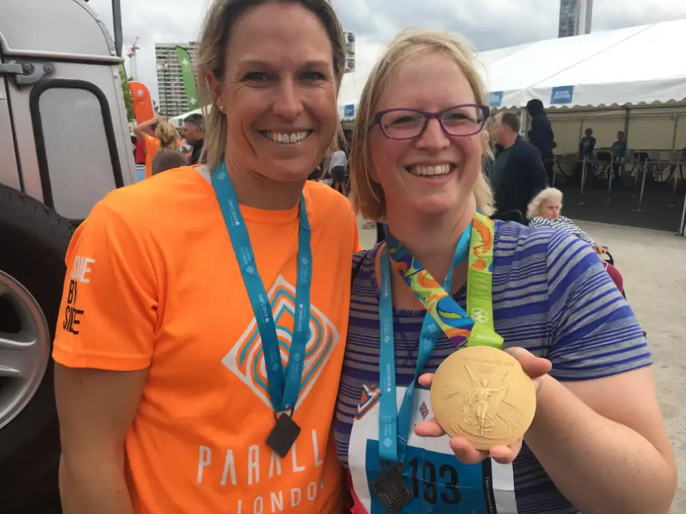 Two women wearing medals smile at an outdoor event; one woman, in a striped shirt, holds a large gold medal.