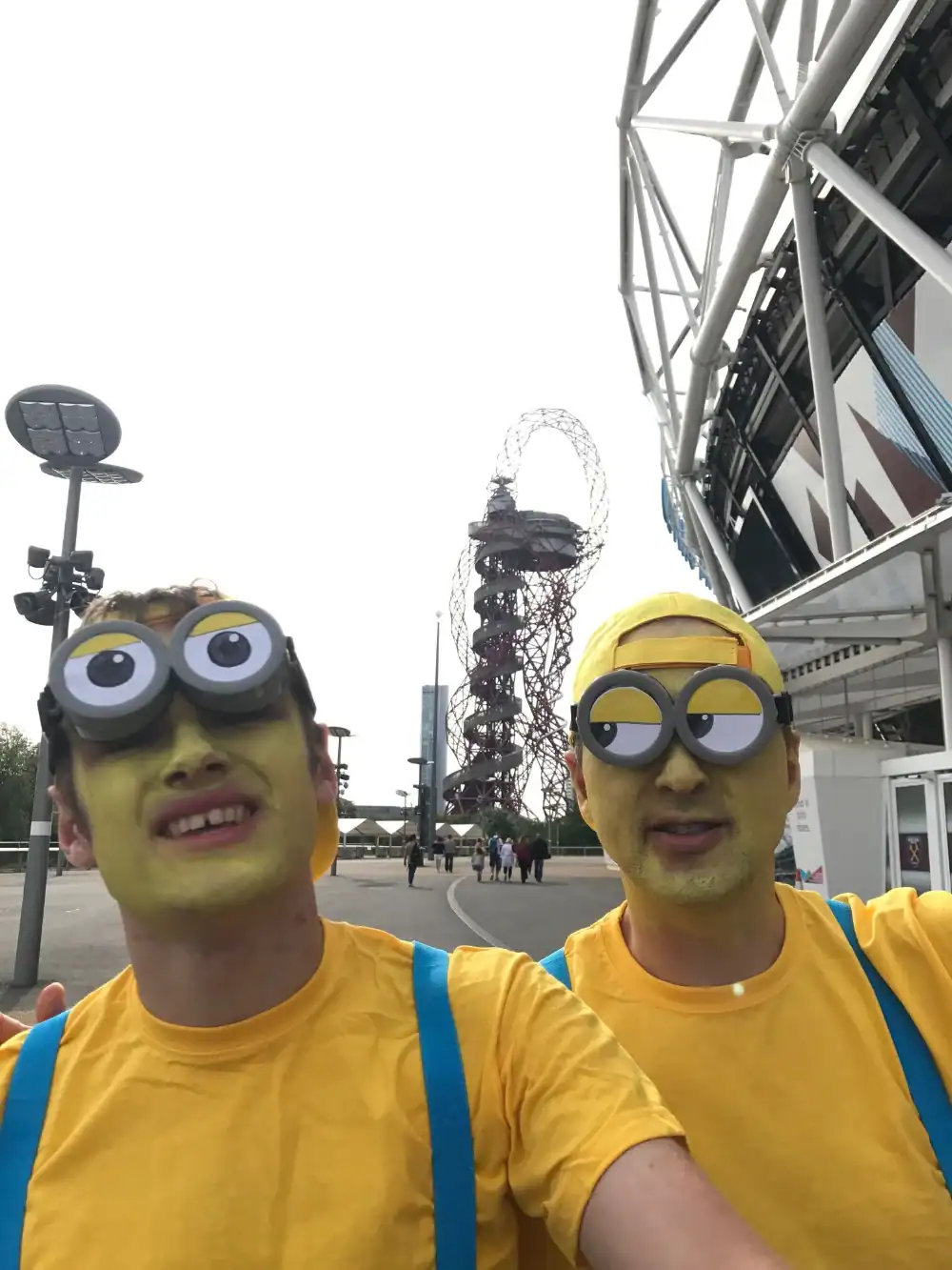 Two people wearing yellow shirts, yellow face paint, yellow hats, blue suspenders, and large Minion-style glasses stand outside near a modern structure and the ArcelorMittal Orbit tower.