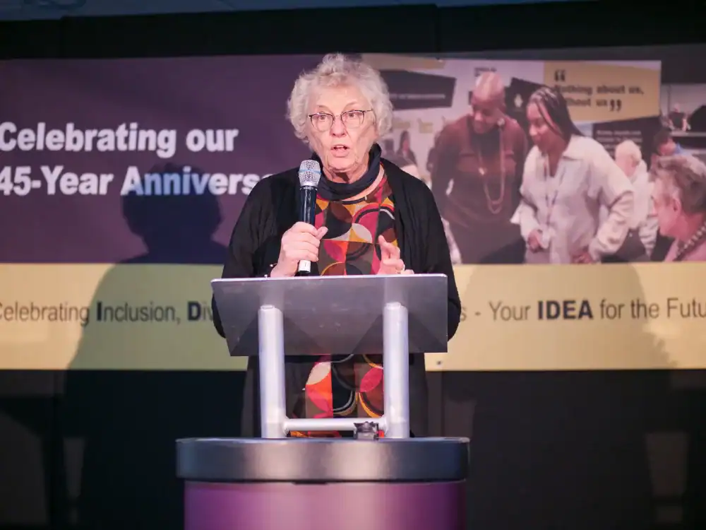 An older woman with gray hair and glasses speaks at a podium during an event celebrating a 45-year anniversary, with a banner in the background.