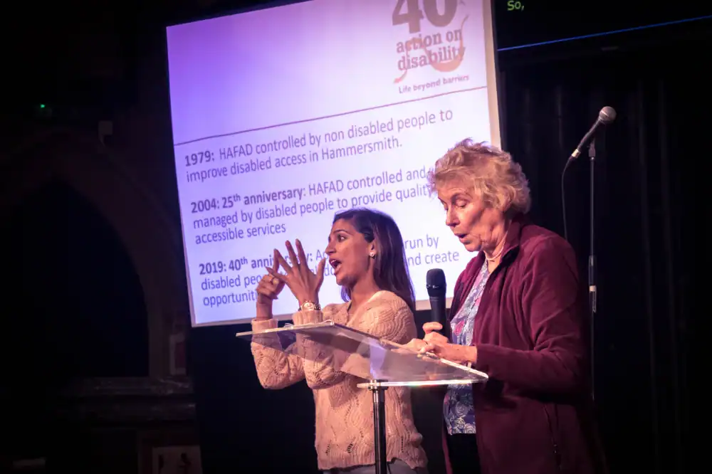 Two women speak on stage; one uses sign language. A projected presentation behind them displays milestones for Action on Disability’s 40th anniversary event.