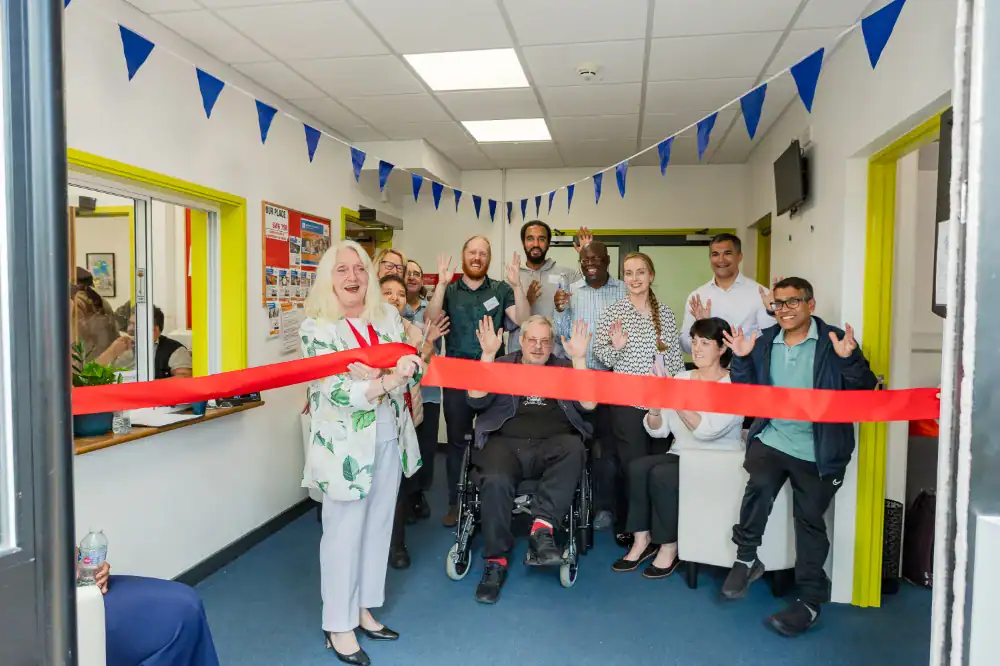 A group of people, some standing and one in a wheelchair, gather and smile as a woman cuts a red ribbon at an indoor event decorated with blue bunting.