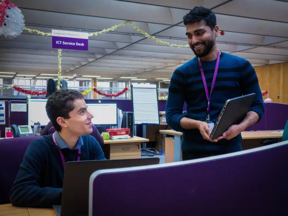 Two men in an office setting, one sitting at a computer and the other standing with a laptop, conversing near an "ICT Service Desk" sign.