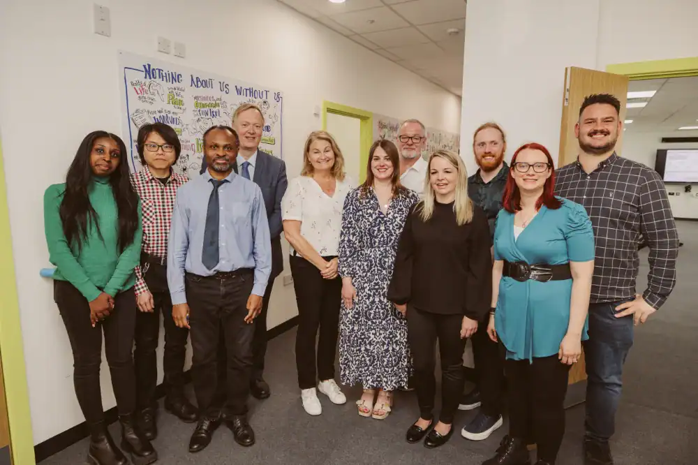 A group of eleven people stand indoors in a hallway, posing for a group photo in front of colorful posters on the wall.