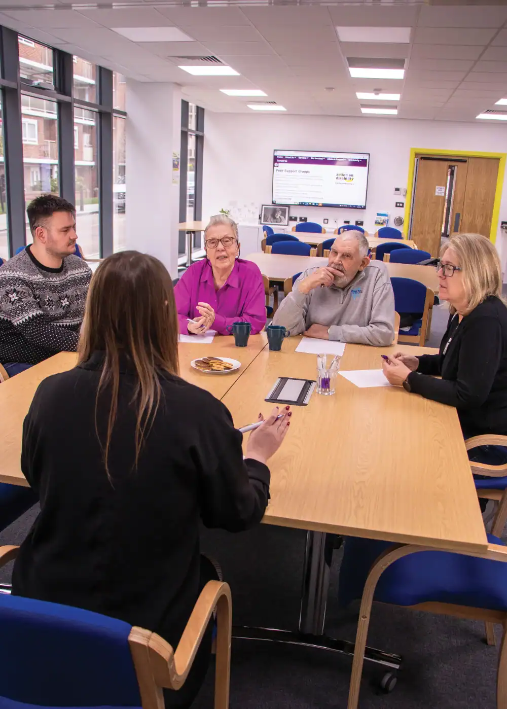 Five people sit around a table having a discussion in a well-lit meeting room with large windows and a screen displaying information in the background.