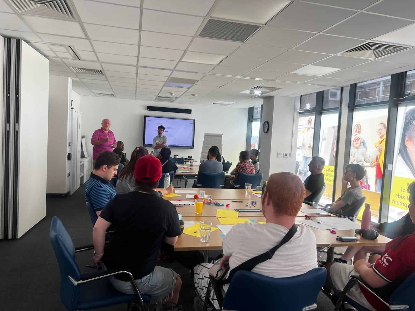A group of people sit around tables in a meeting room, listening to two presenters standing near a screen and flip chart at the front of the room.