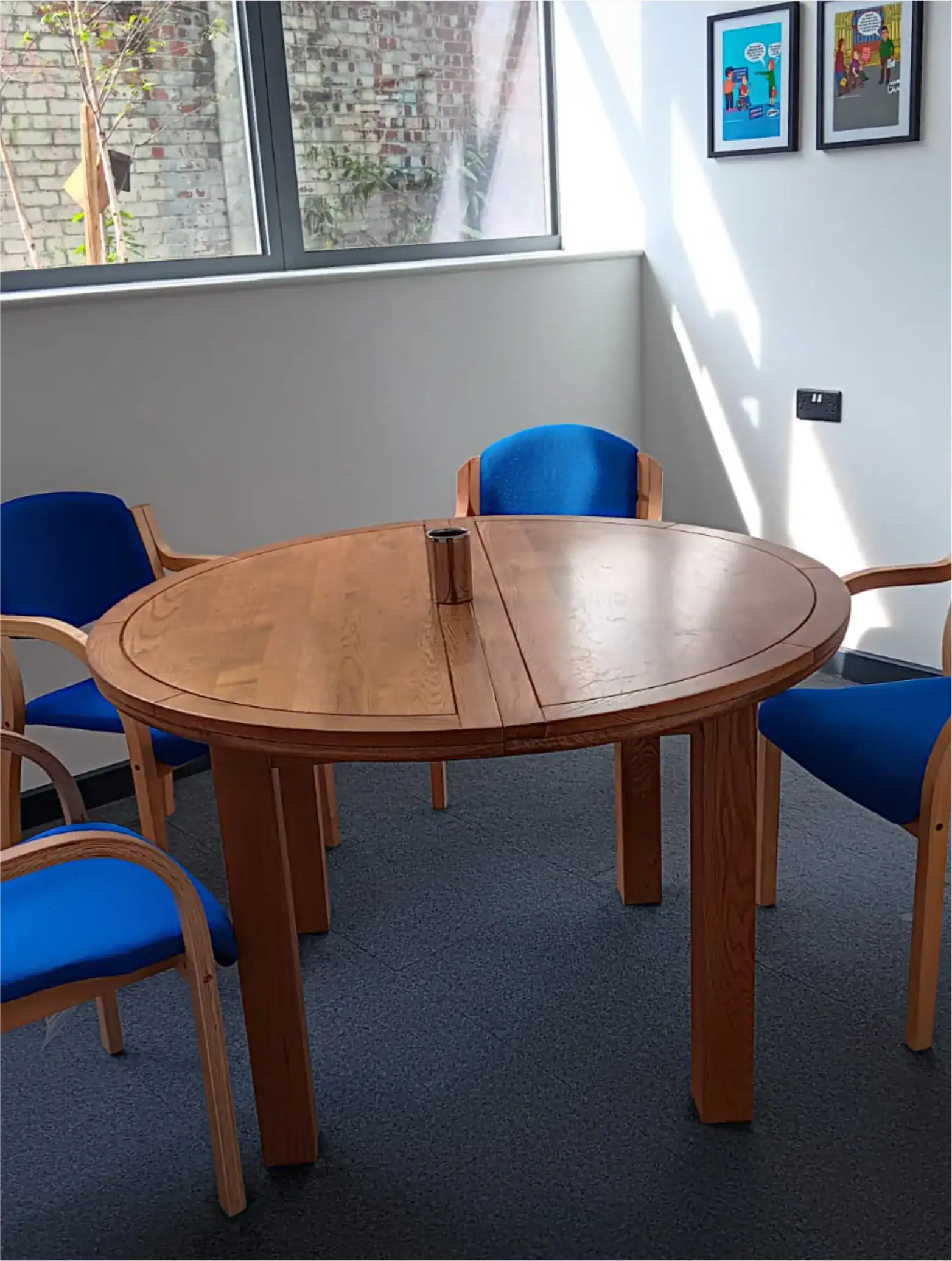 A round wooden table with a split down the middle is surrounded by four blue and wood chairs in a sunlit room with framed pictures on the wall and a window facing a brick wall.