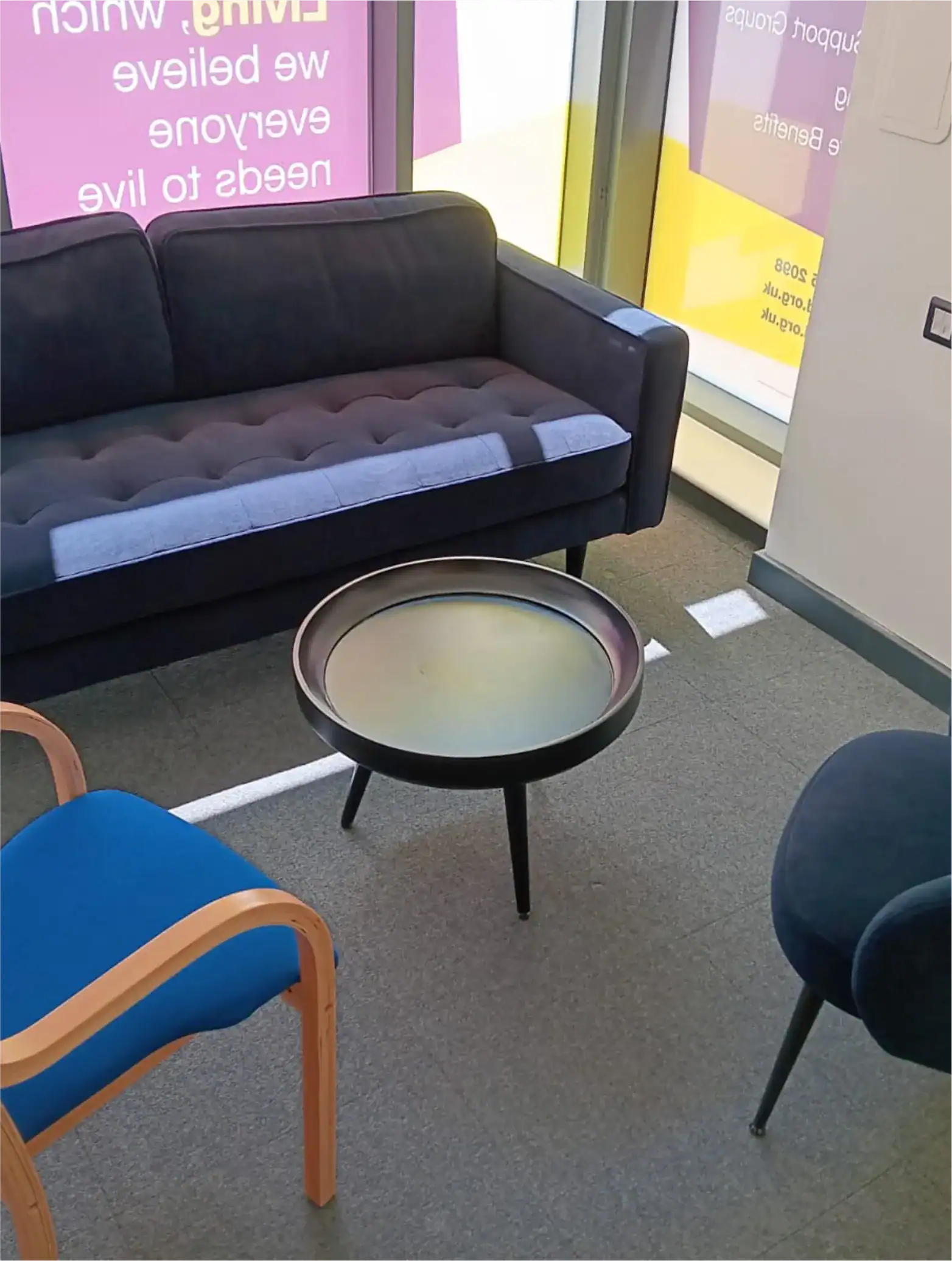 A small waiting area with a dark sofa, two chairs, and a round black coffee table on a gray carpeted floor, near large windows with pink and yellow signs outside.