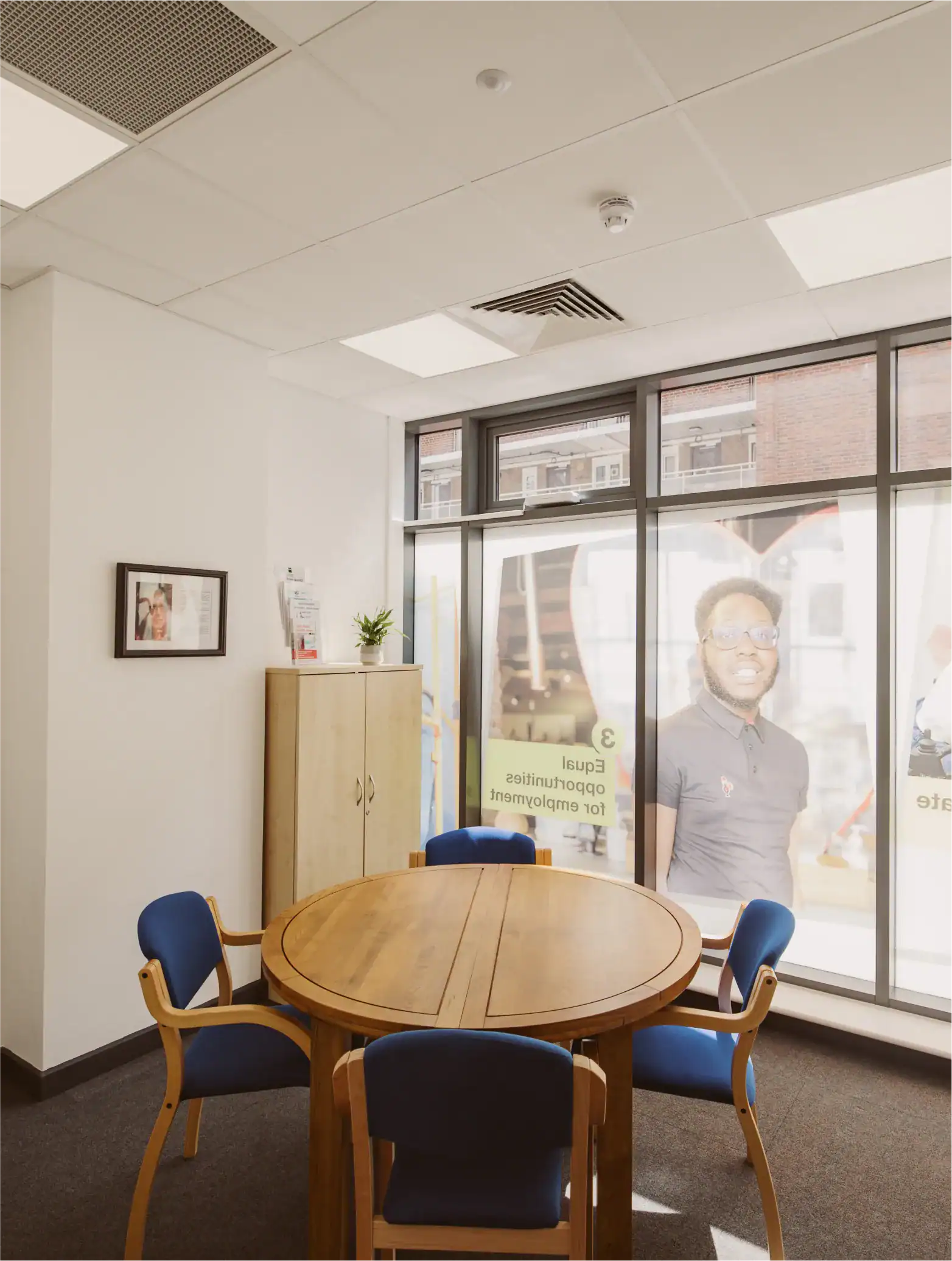 A small office meeting room with a round wooden table, four blue chairs, a wooden cabinet, and large windows displaying a poster of a smiling man outside.