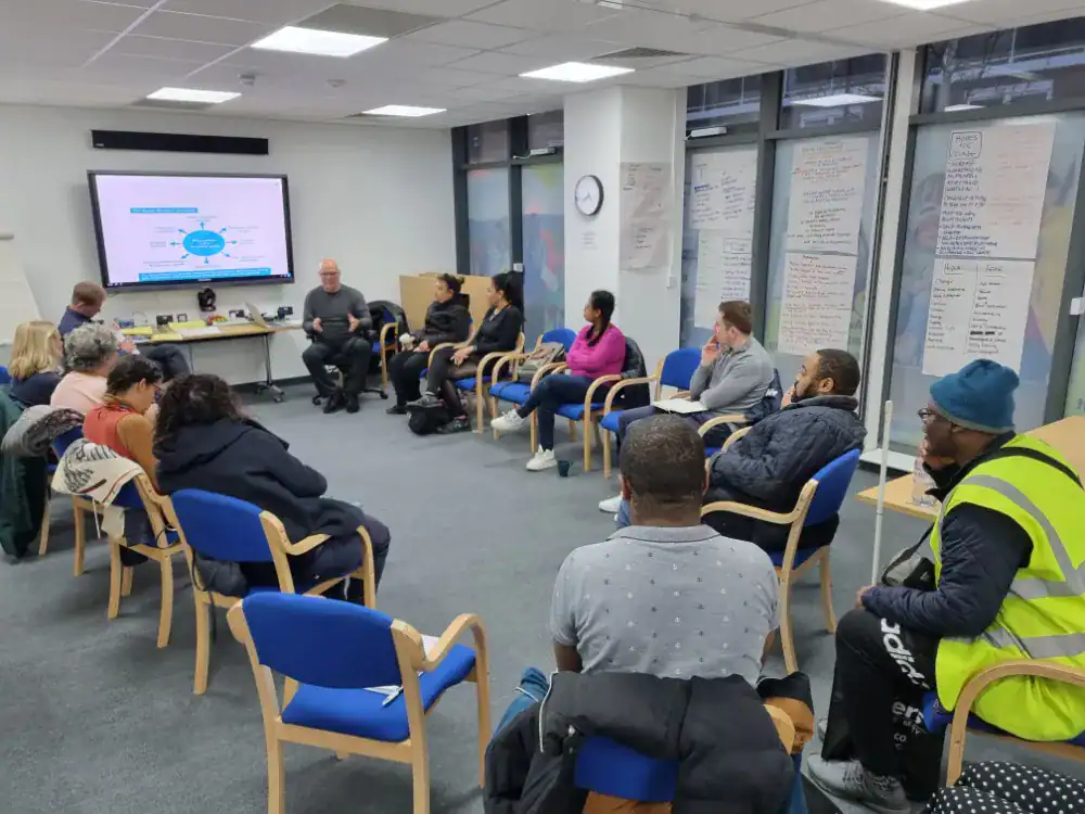 A group of people sit in a circle in a meeting room, listening to a speaker near a large display screen showing a presentation.